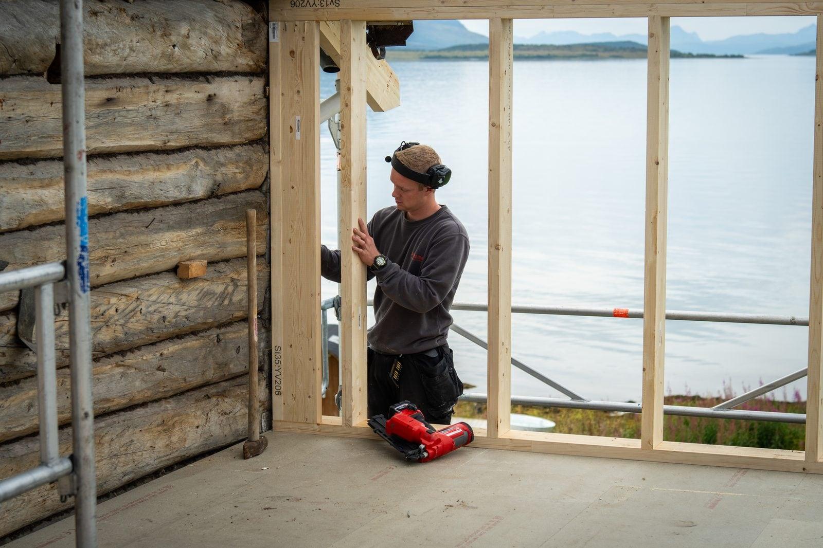 Tømrer i arbeid i et tømmerhus, med utsikt mot sjøen